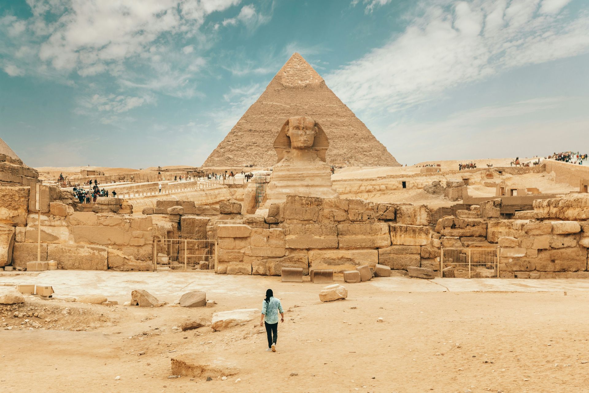 A woman is walking in front of a pyramid and sphinx in the desert.
