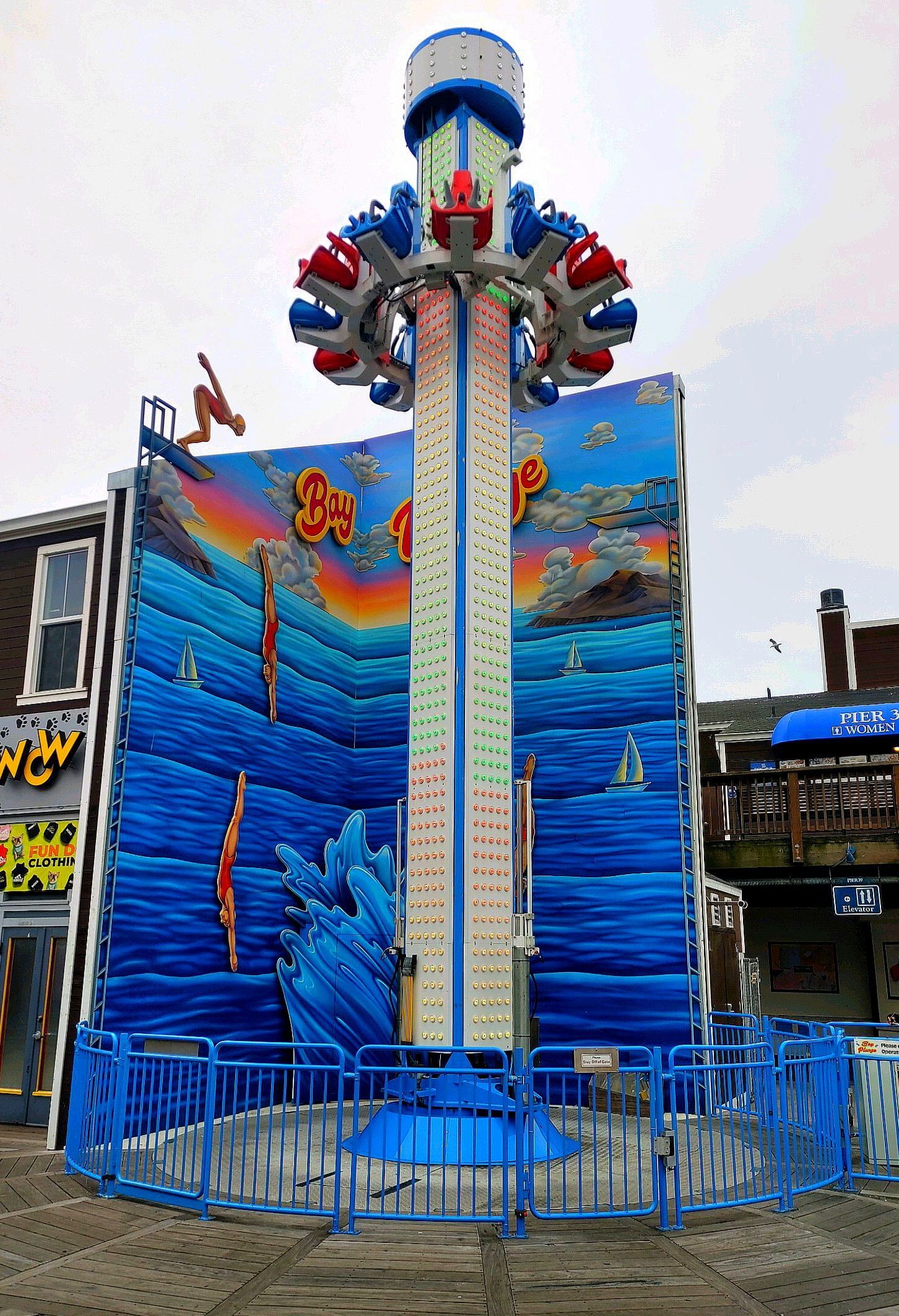 Amusement ride tower with blue mural panels and red-and-blue seats enclosed by blue fencing