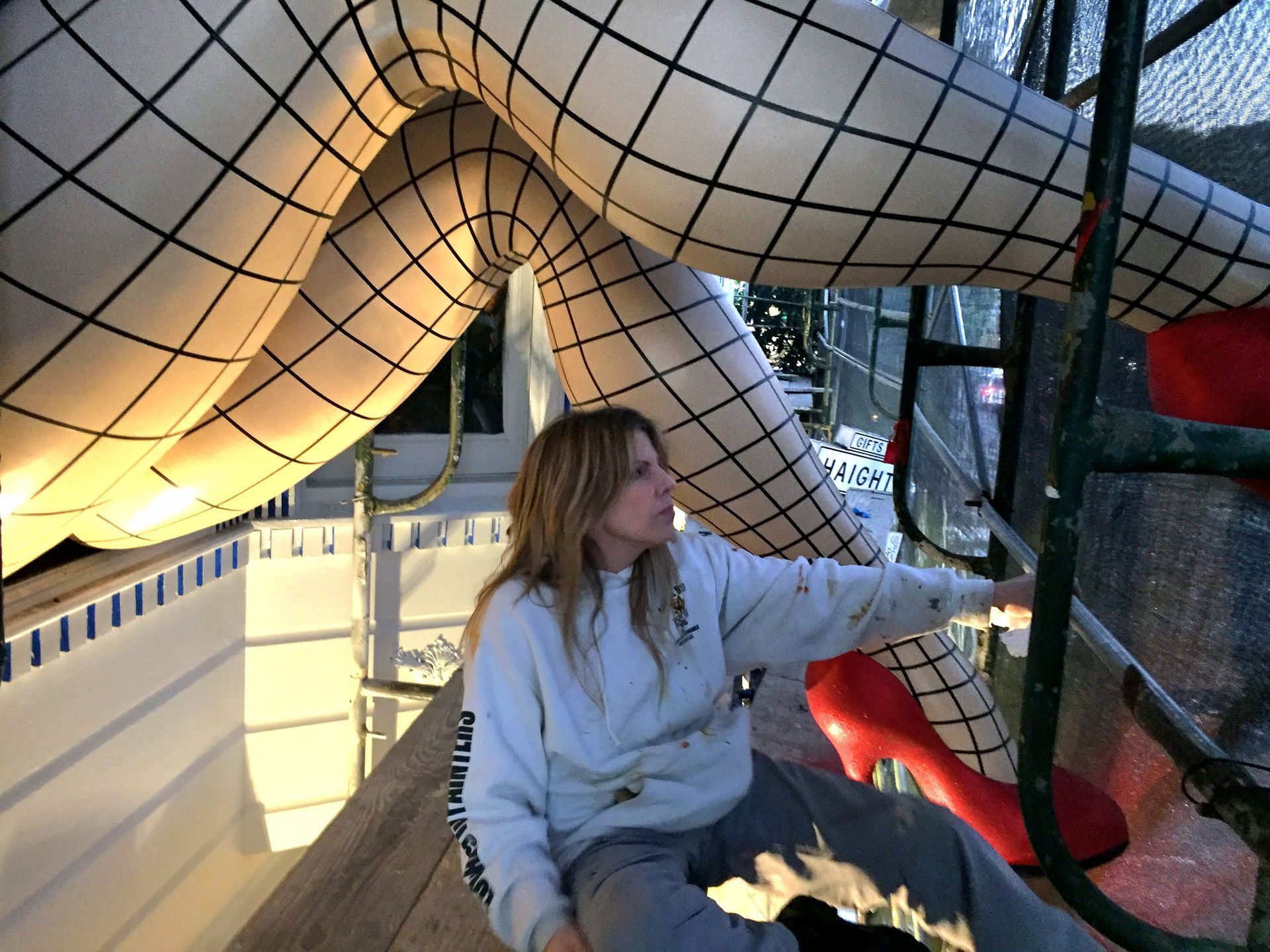 Person in a gray hoodie seated under a large netted sculpture, reaching toward a red-lit opening.