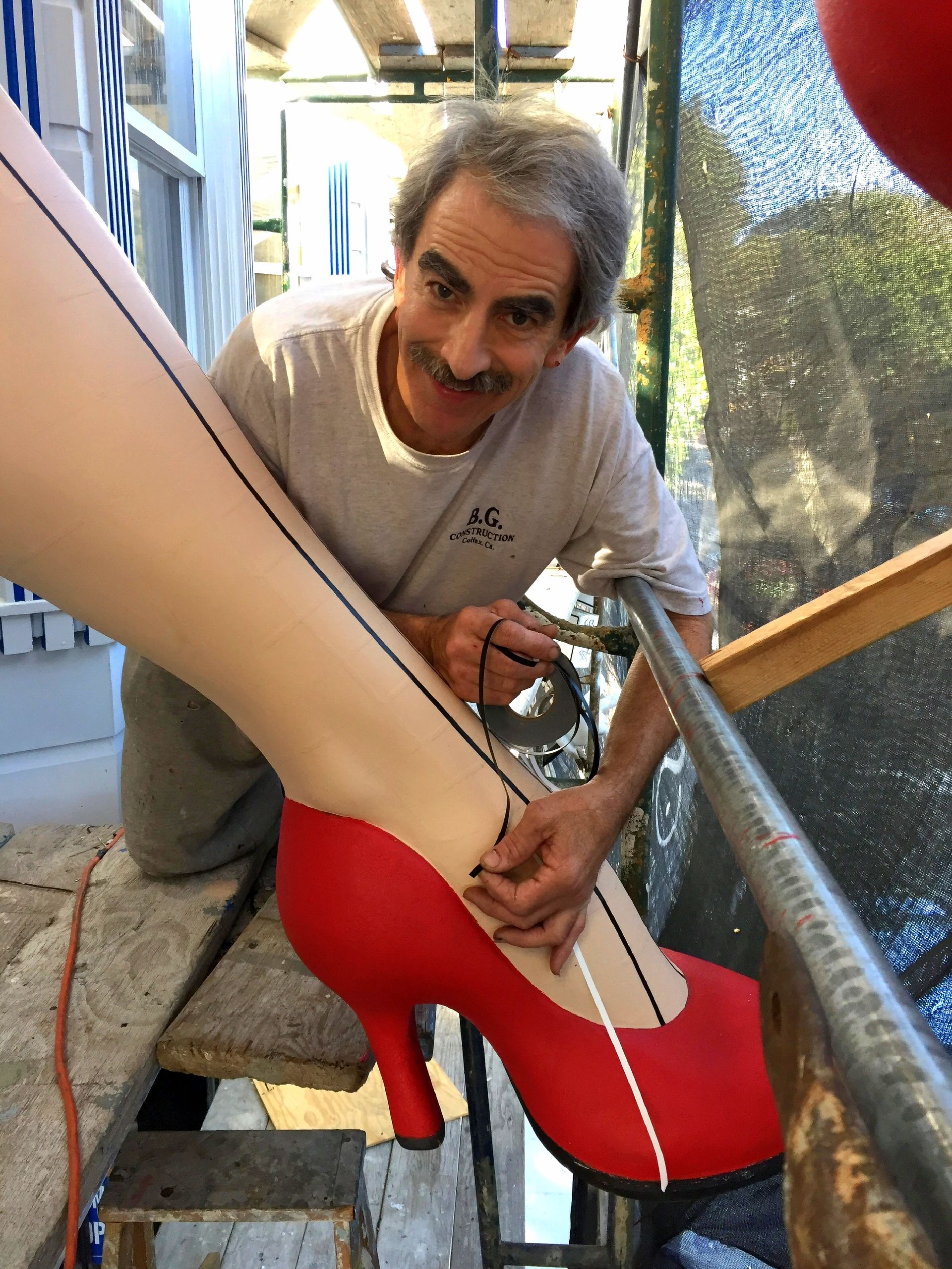 Person smiling and working on a red high-heeled shoe under a large mannequin leg in a workshop