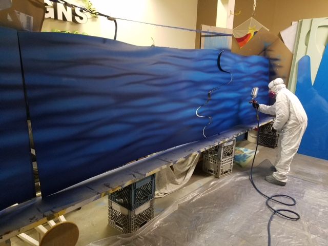 Person in white suit spray-painting a large blue mural on a wall in a workshop