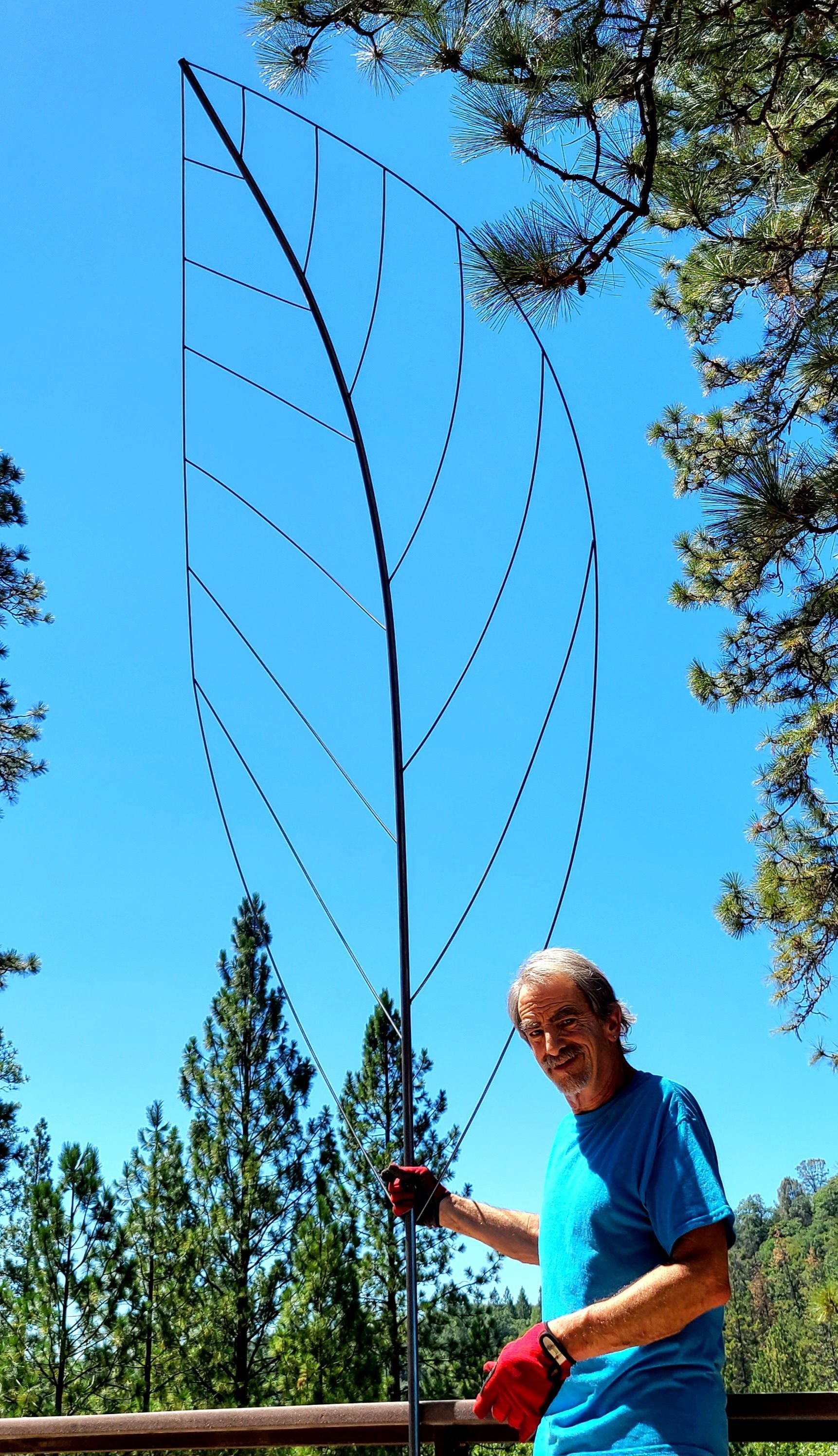 Person in a blue shirt and red gloves holds a tall wire sculpture outdoors against a blue sky.