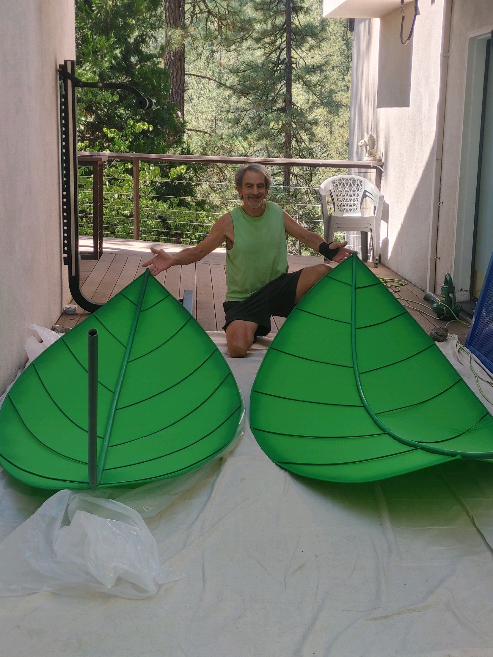 Child kneeling between two large green leaf-shaped crafts on a balcony or patio