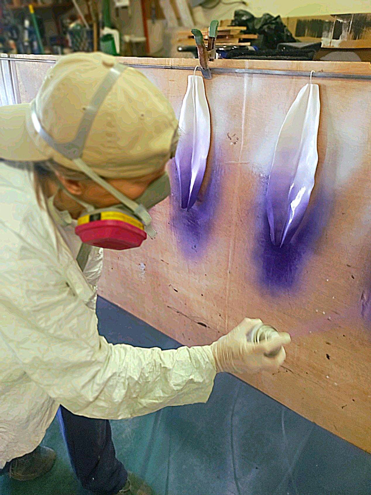 Person in protective gear spraying purple paint onto a wooden panel in a workshop