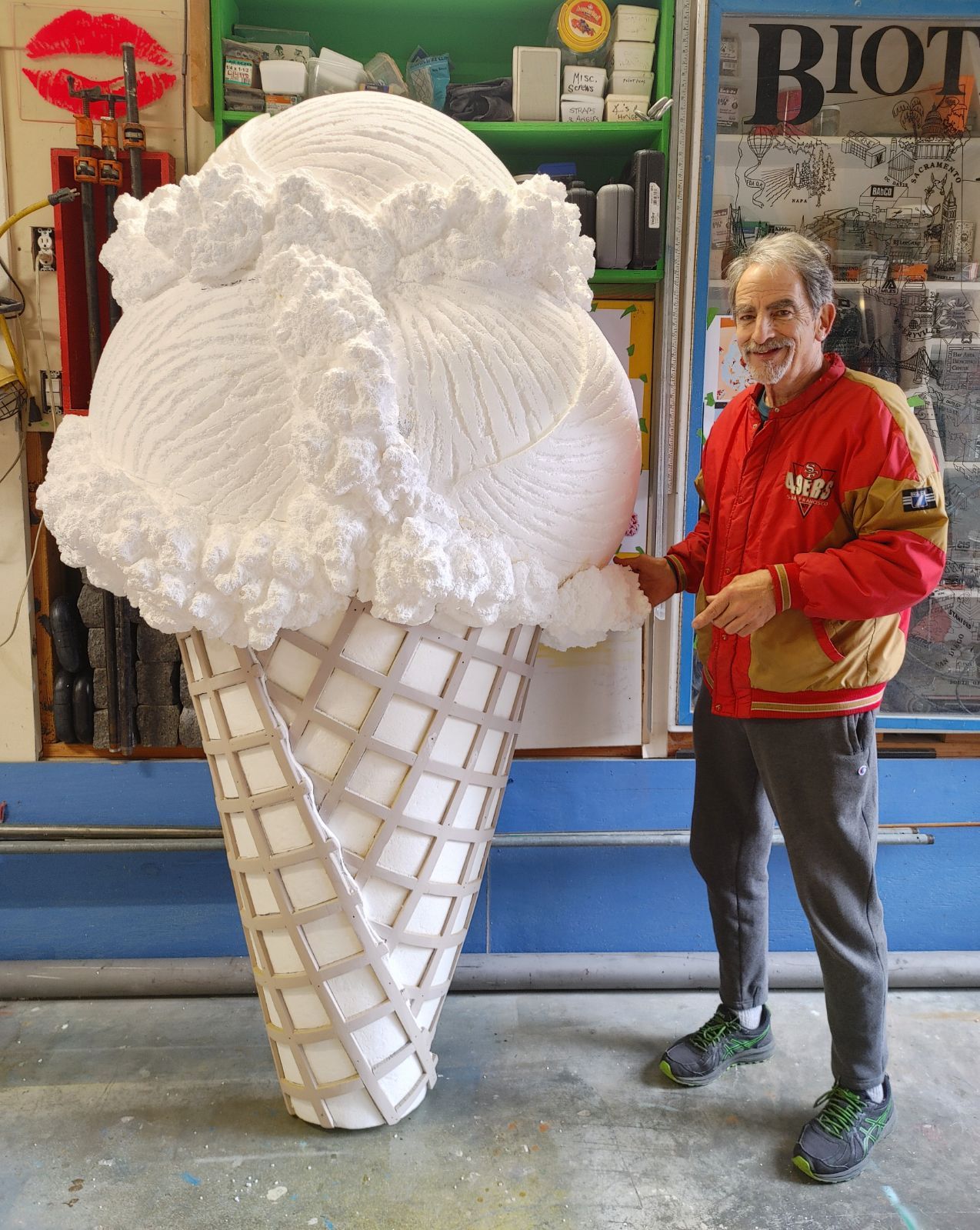 Man standing beside a giant white ice cream cone sculpture in a workshop