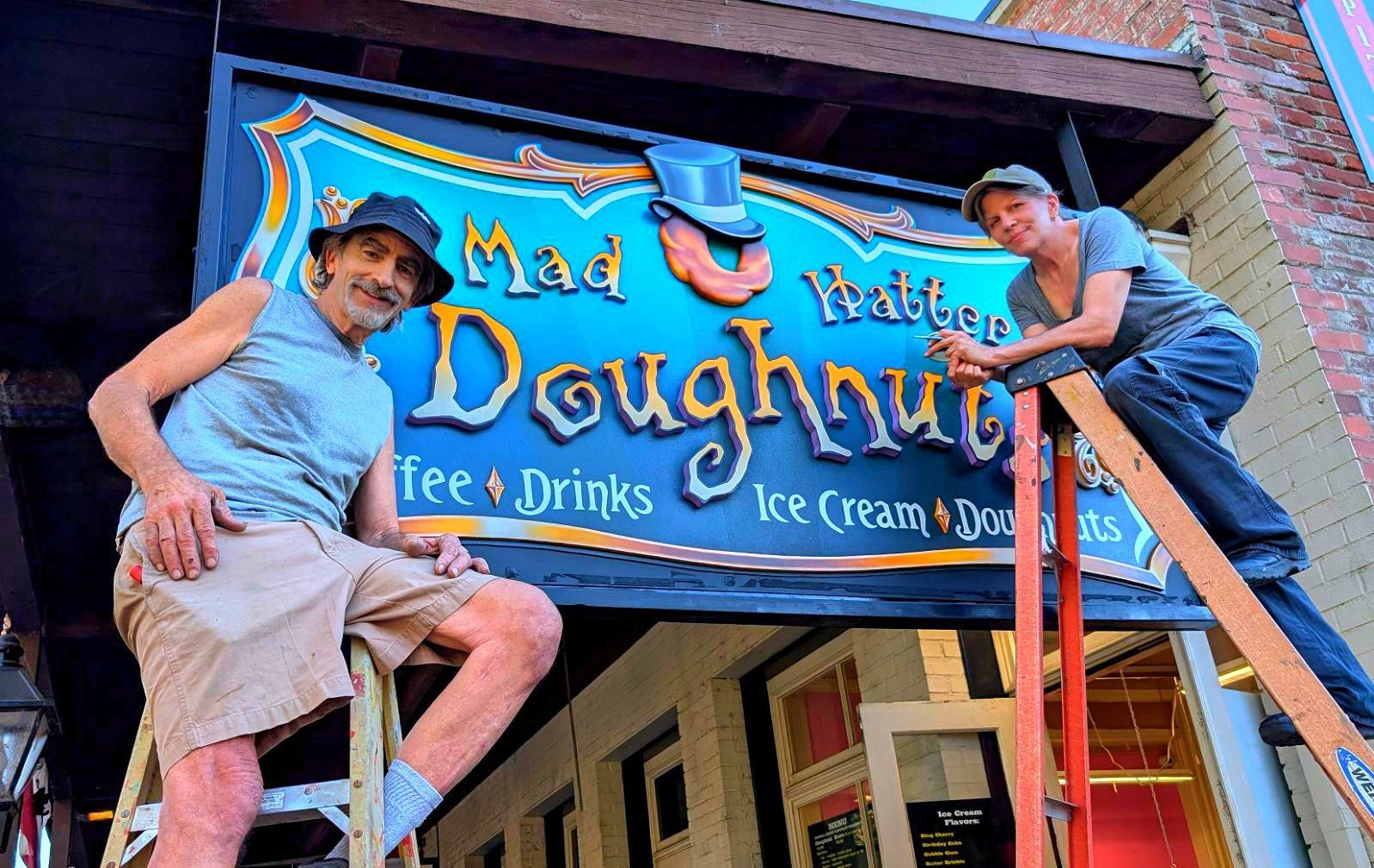Two workers on ladders painting a bright blue ice cream shop sign reading “Mad Hatter Doughnuts.”