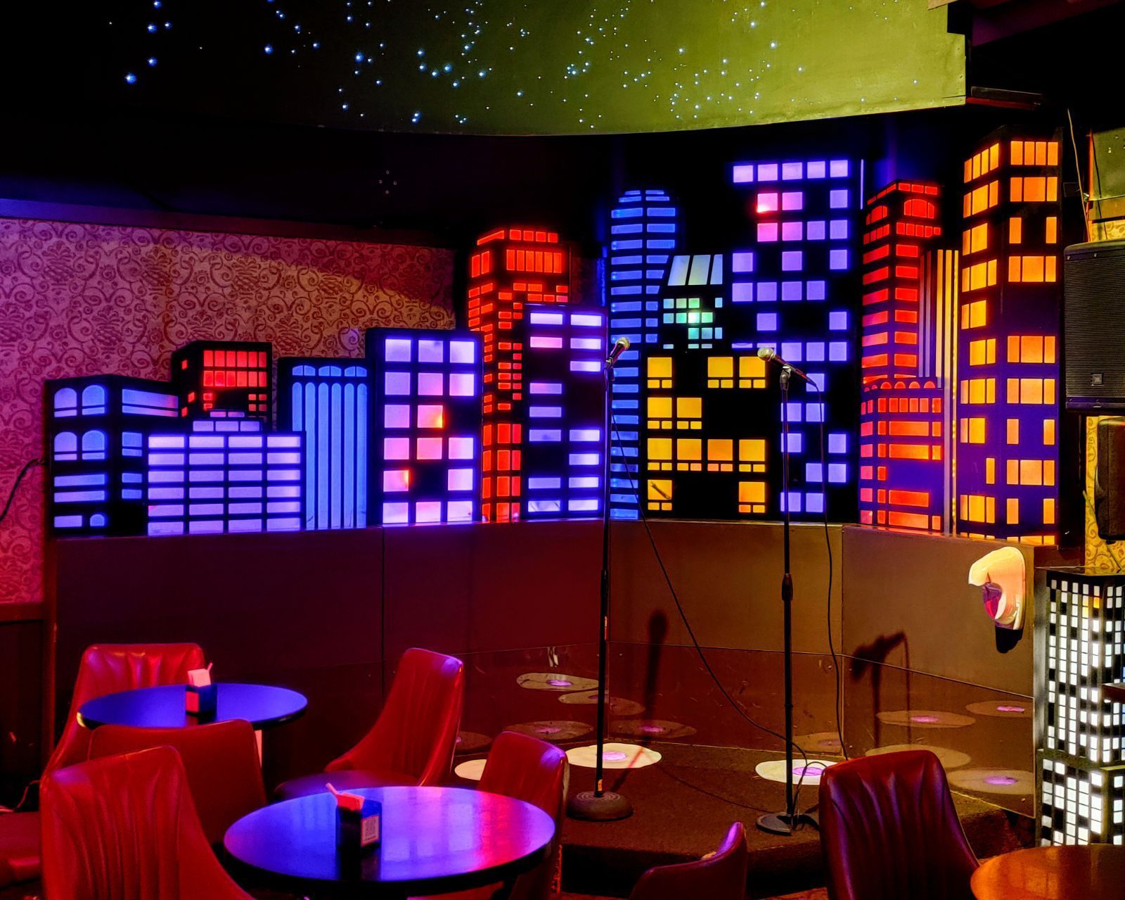 Neon-lit lounge with city skyline mural, red chairs, and small round tables under colorful lights
