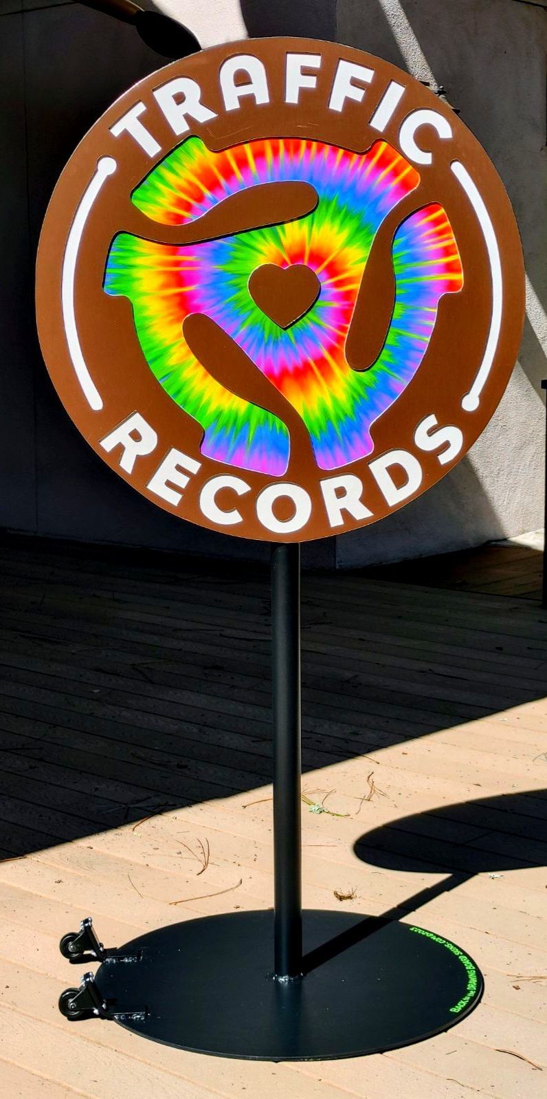 Round “TRAFFIC RECORDS” sign with rainbow tie-dye logo on a black post outdoors, casting a shadow
