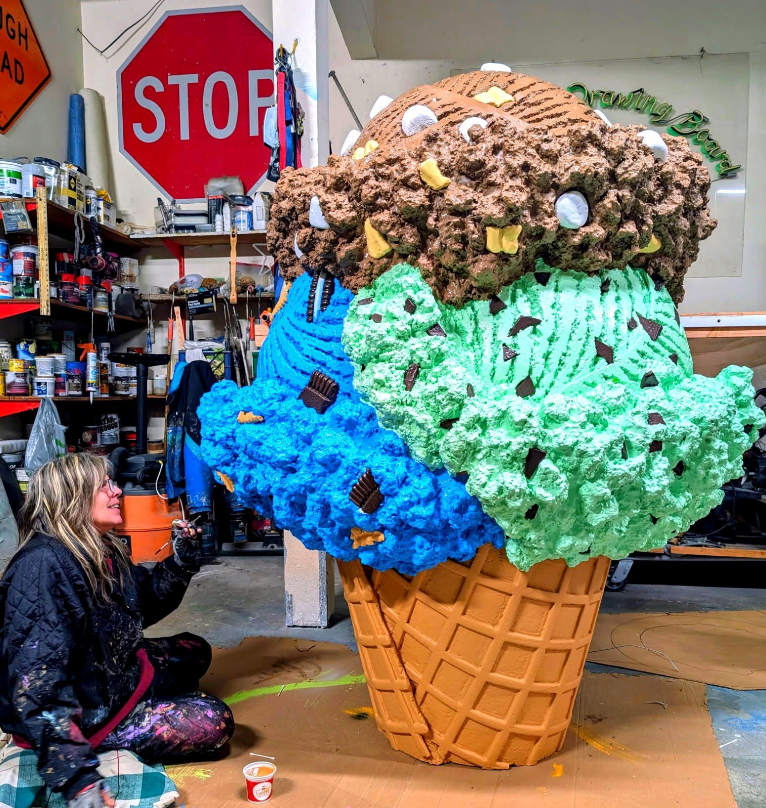 Giant ice cream cone sculpture with blue, green, and brown scoops in a shop, beside a seated person and stop sign