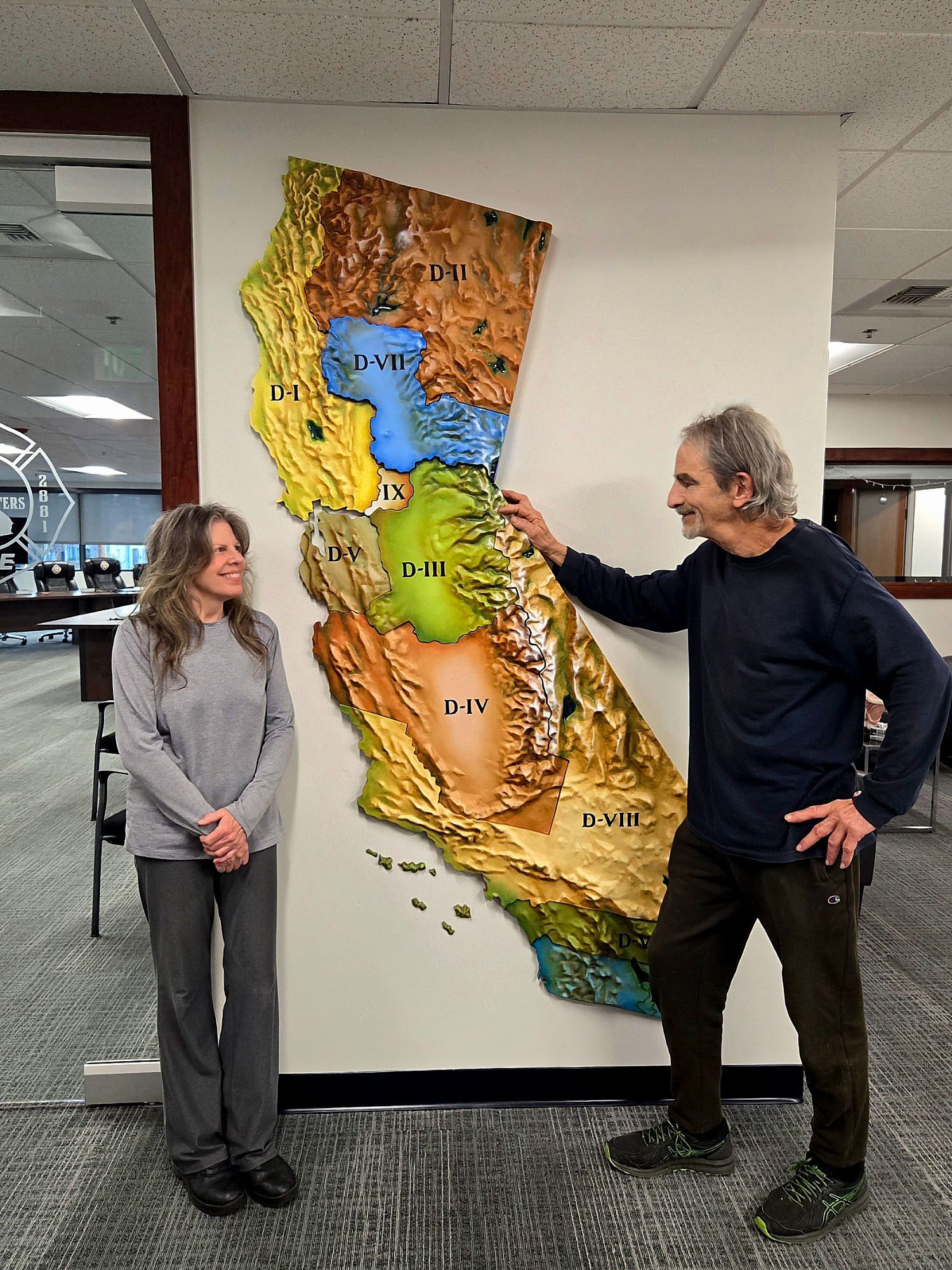 Two people stand beside a colorful wall relief map, one pointing at a blue lake area.
