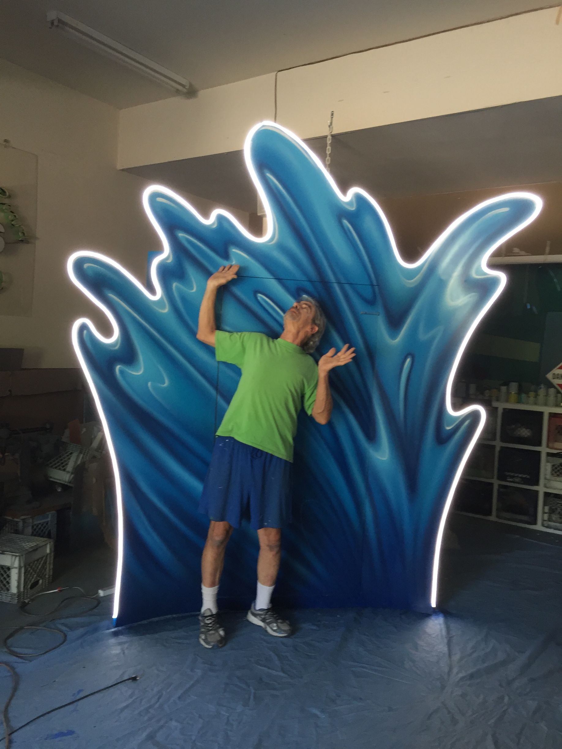 Person posing beside a blue wave-shaped art piece indoors, wearing a green shirt and black shorts.