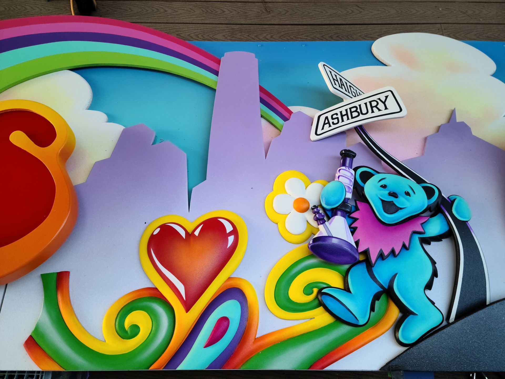 Colorful cartoon scene with rainbow, heart, clouds, and a blue character holding an Ashbury sign