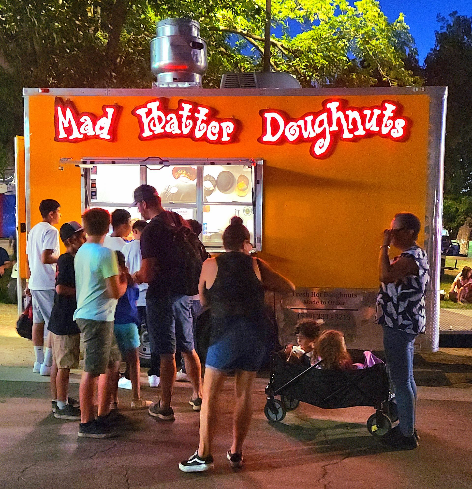 People waiting at the Mad Hatter Doughnuts truck at dusk, with a lit service window and neon sign.