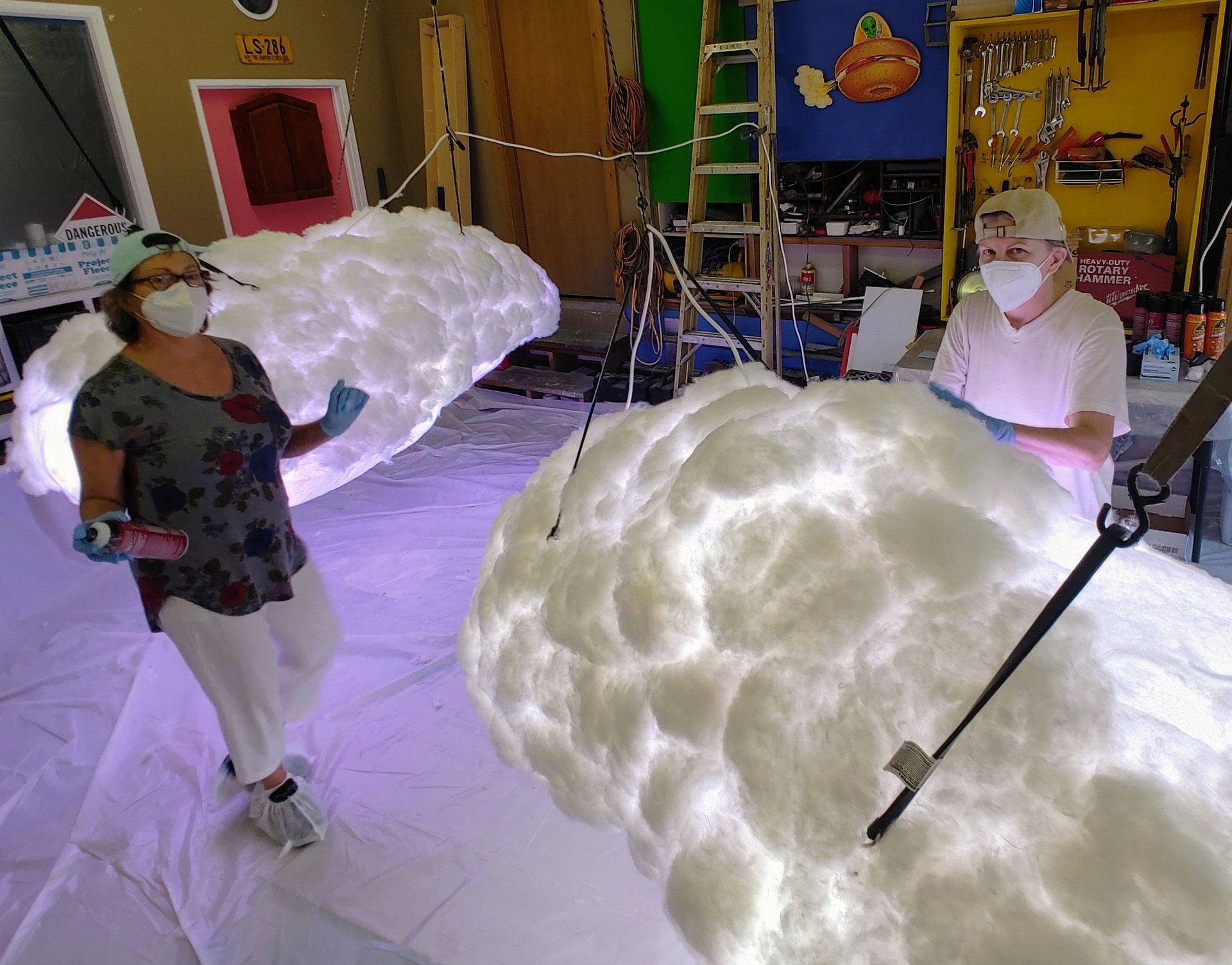 Two masked children stand by glowing cloud-shaped props in a colorful classroom.