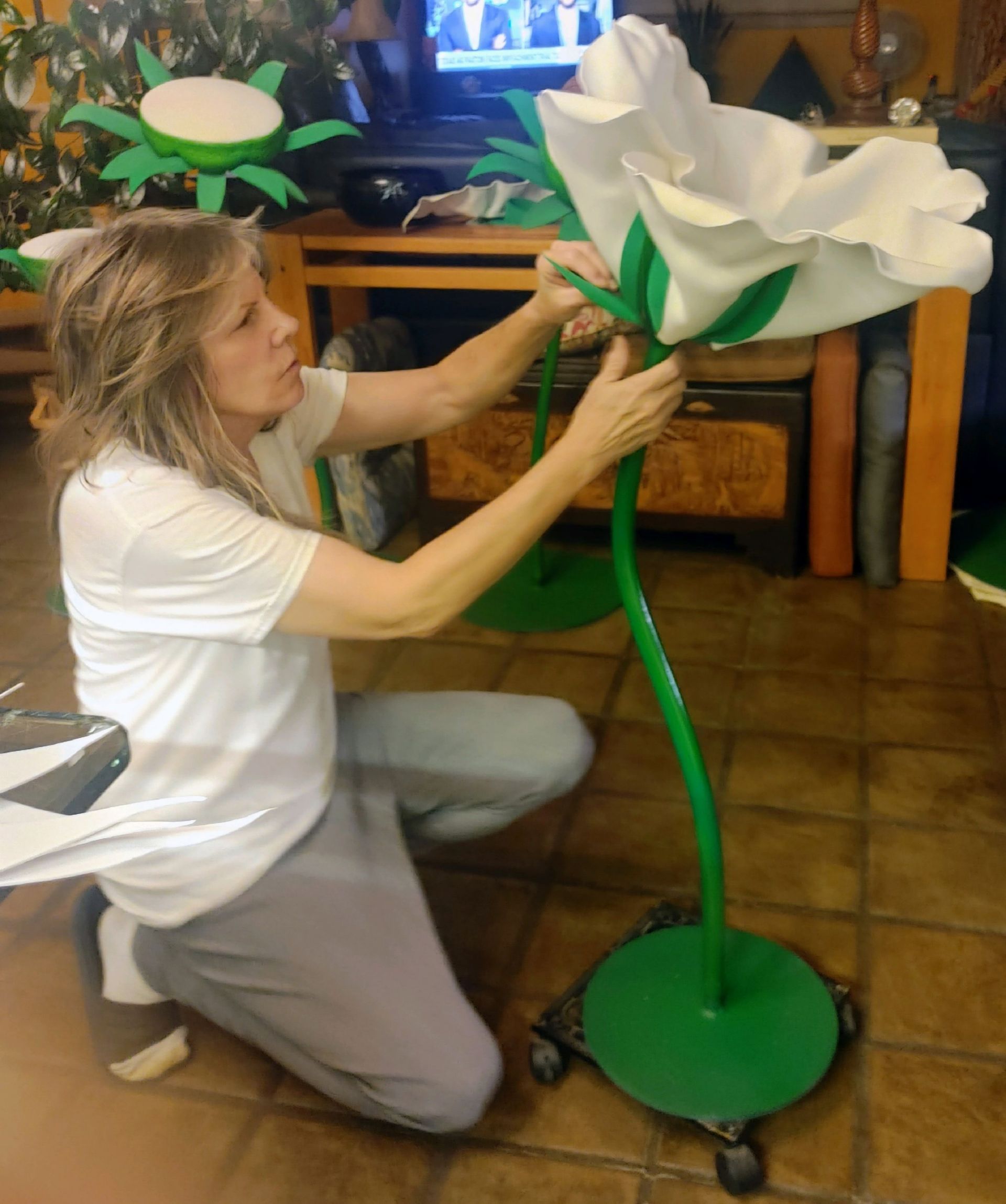 Woman kneeling, adjusting a large white flower sculpture with green stem and base indoors.