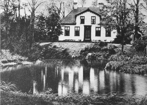 Een klein wit huisje weerspiegeld in een vijver, omgeven door kale bomen en gras.