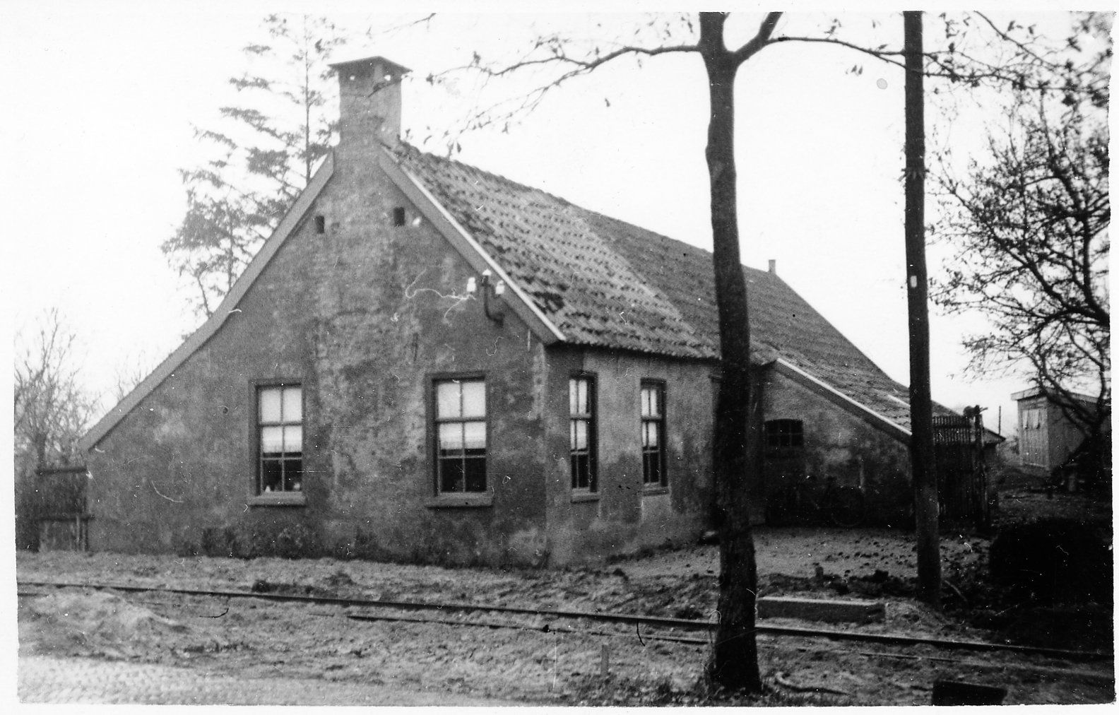 Zwart-witfoto van een klein bakstenen huis met een pannendak en kale bomen in de tuin.