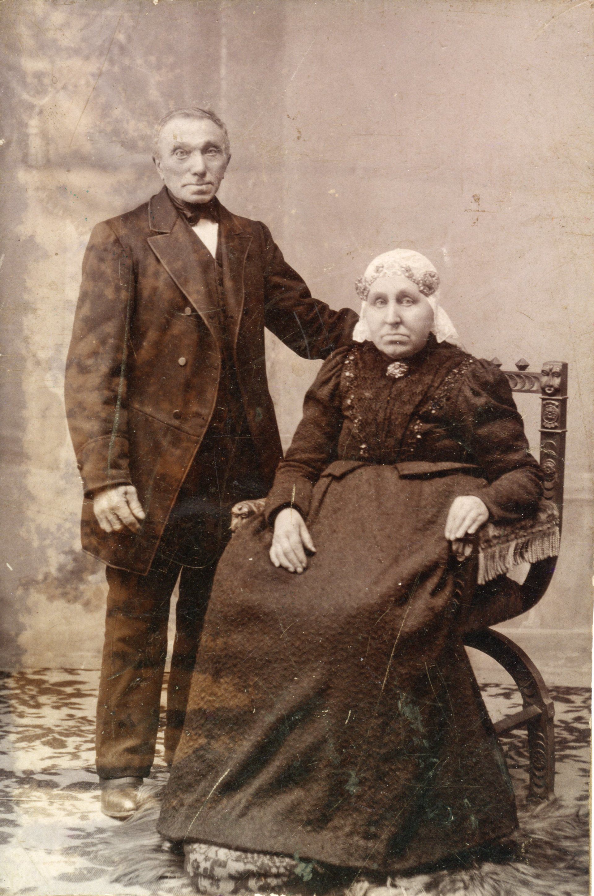 Sepia studiofoto van een staande man naast een zittende vrouw in een donkere jurk en op een stoel.