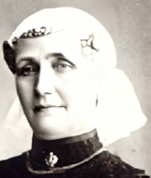 Zwart-witportret van een persoon met ceremoniële hoofdtooi en donkere kleding met hoge kraag, die iets naar links kijkt.