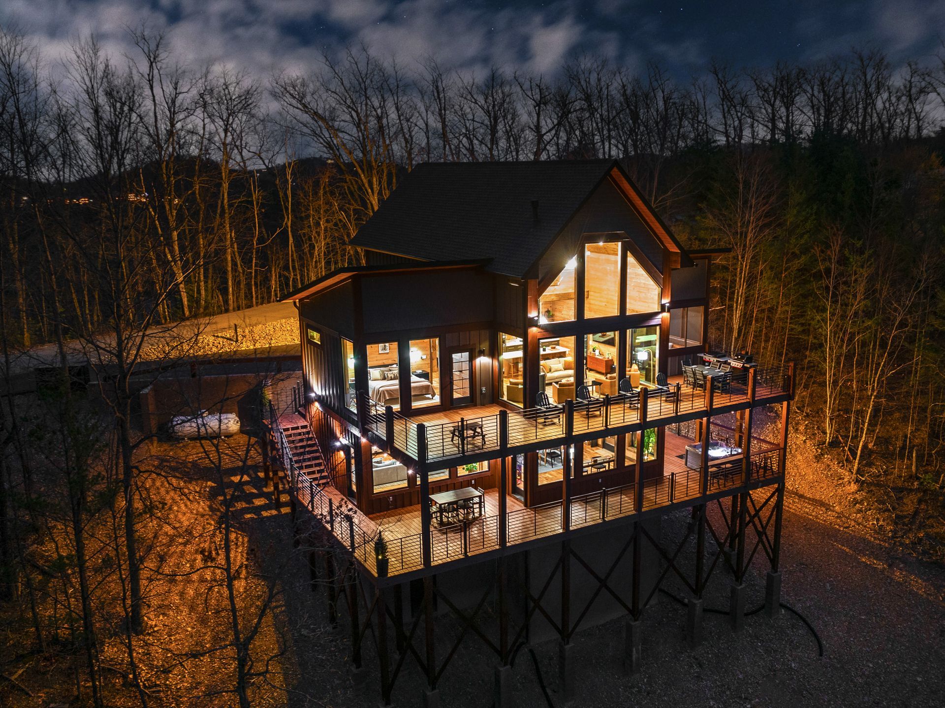 A modern cabin on stilts illuminated at night, nestled in a dark forest under a starry, cloudy sky.