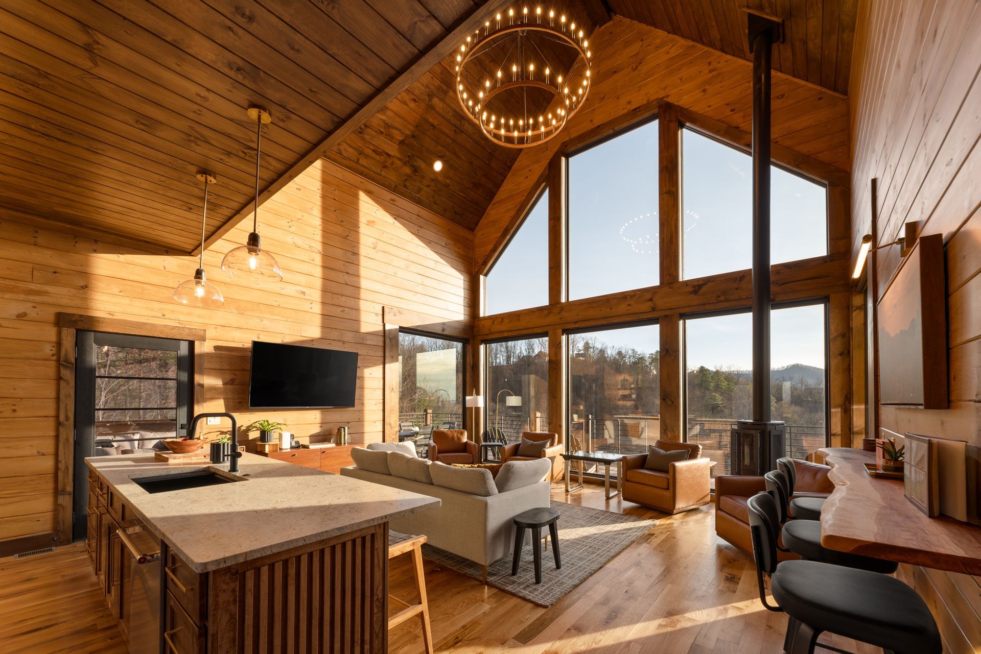 Sunlit wooden cabin interior featuring a vaulted ceiling, kitchen island, living area, and large windows with outdoor views.