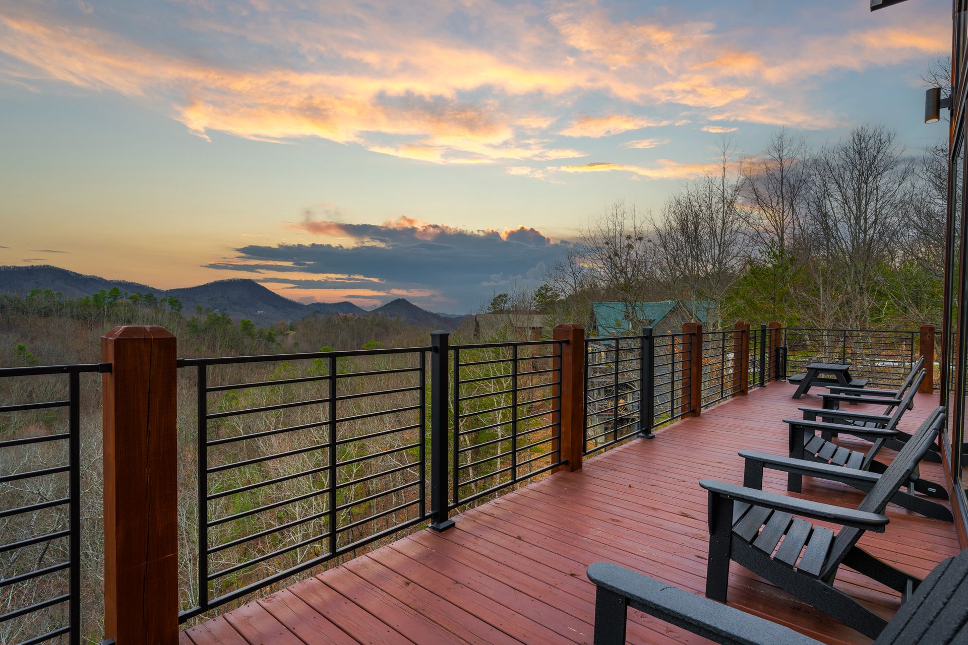 A wooden deck with a black metal railing overlooking a mountain range at sunset with a row of black chairs.