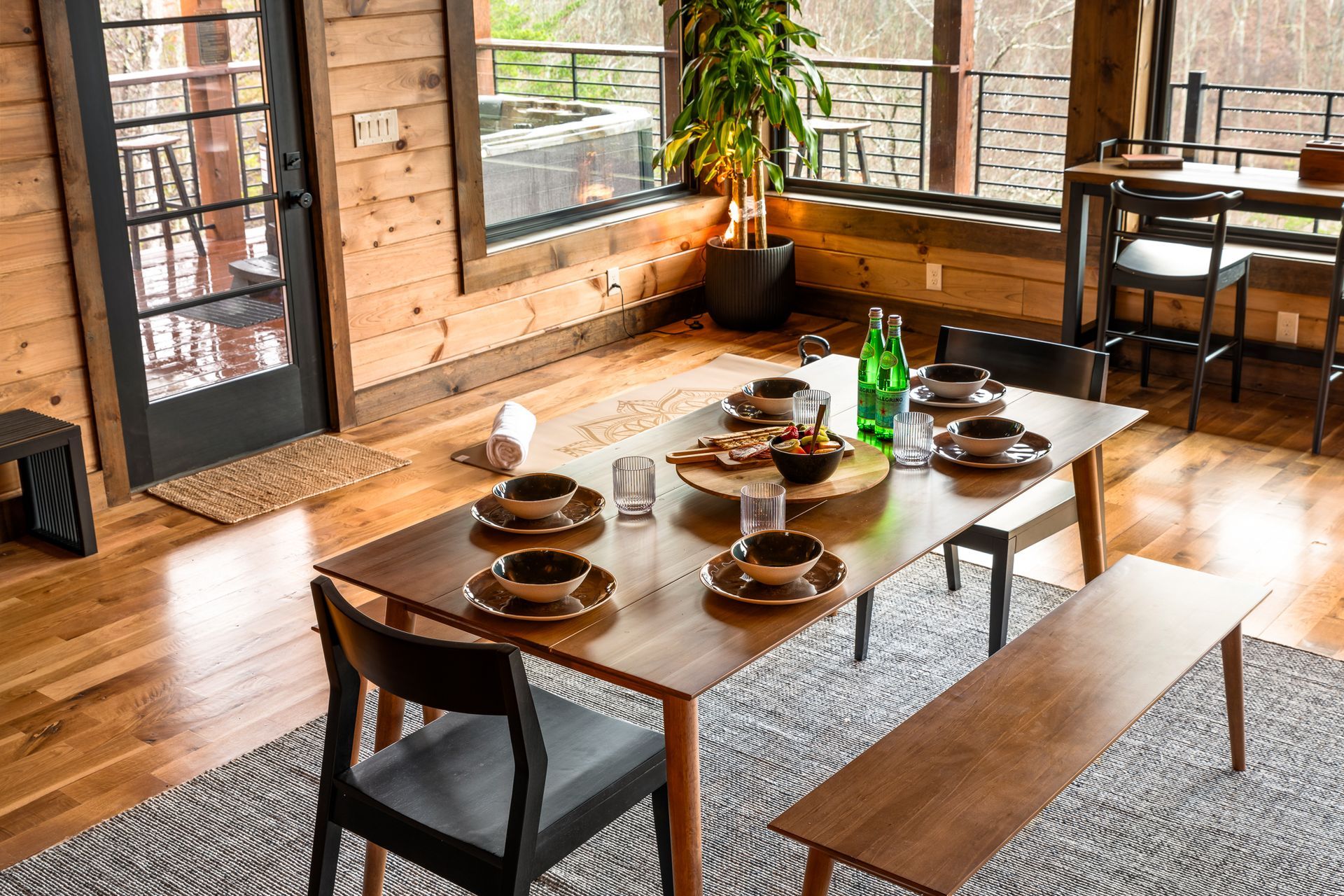 A modern dining set with chairs and a bench sits on a patterned rug in a sunlit wooden room with large windows.