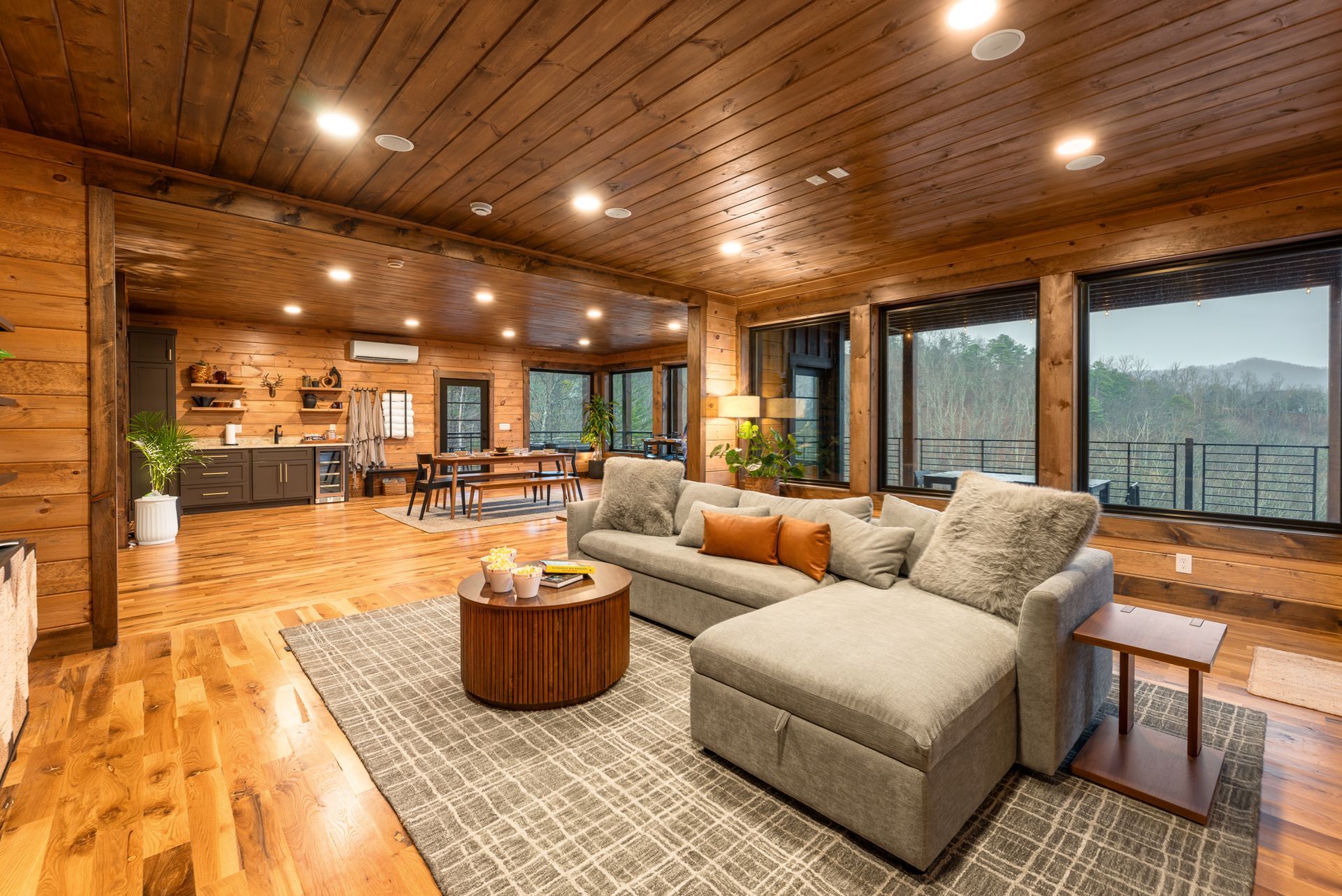 A rustic living room with wood paneled walls, a gray sectional sofa, a round wooden coffee table, and a scenic window view.