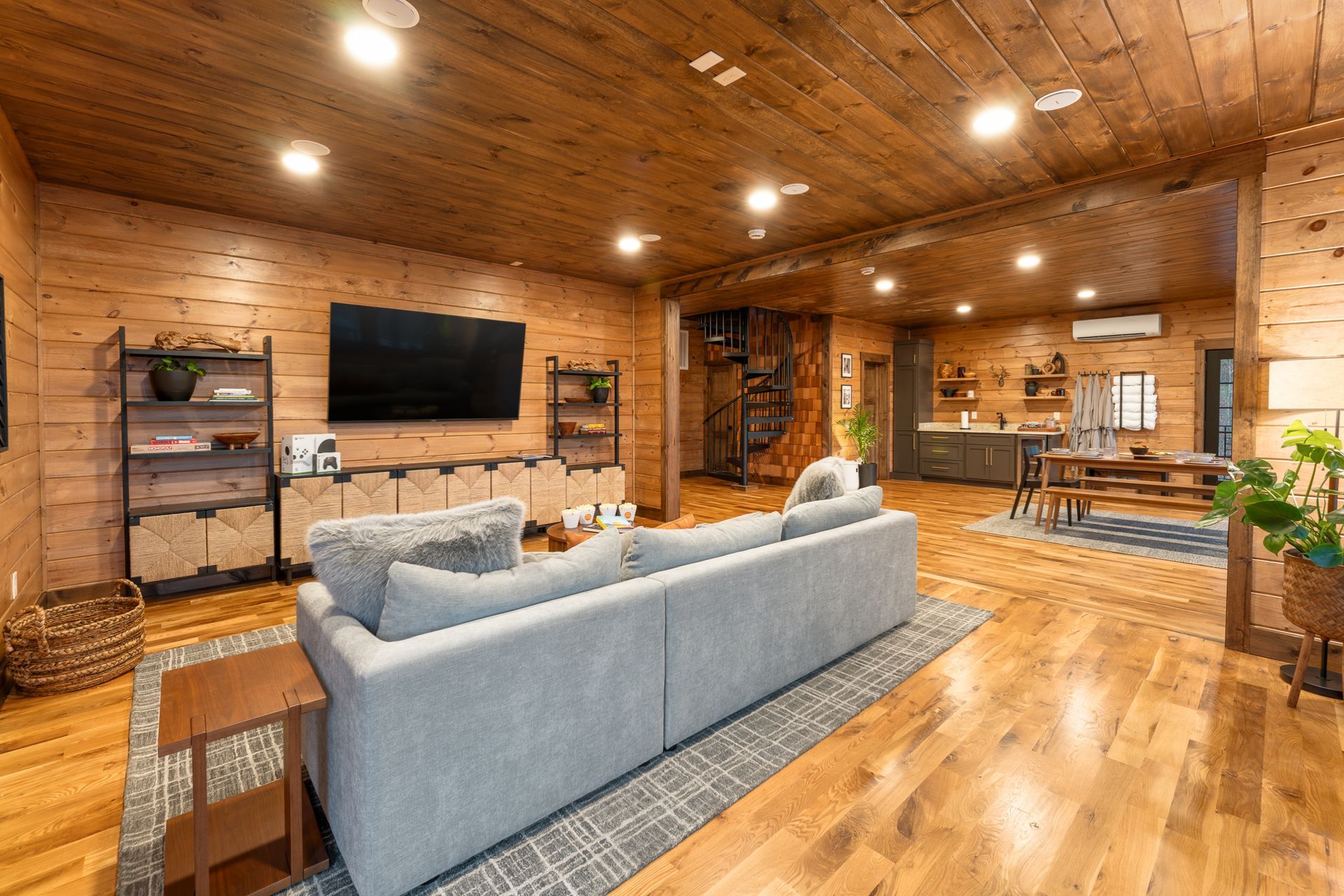 A spacious, rustic log cabin living room with wooden walls and ceiling, a gray sofa, large TV, and an open kitchen area.