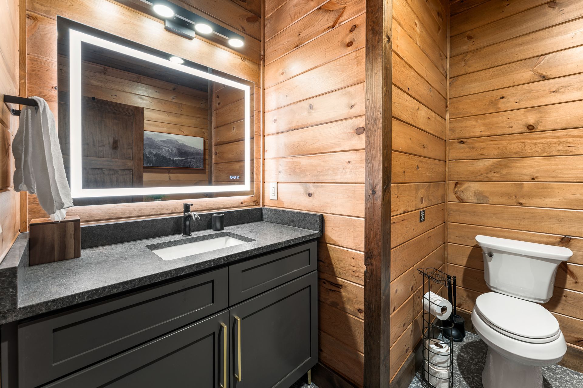 A rustic bathroom with wood walls, a dark grey vanity, a modern illuminated mirror, and a white toilet.