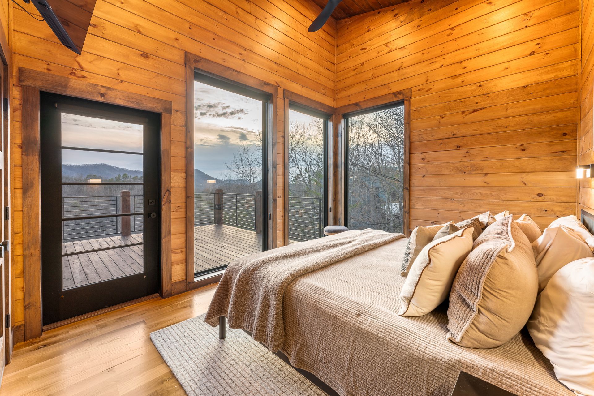 A cozy bedroom with warm wood paneled walls, a large bed with neutral bedding, and floor-to-ceiling windows viewing nature.