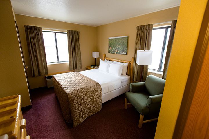 Hotel room with a bed, chair, and two windows with brown curtains. Beige walls and burgundy carpet.