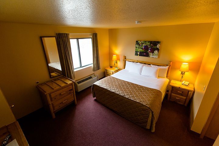 Hotel room with a queen bed, bedside tables, and a dresser. Warm lighting and a window.