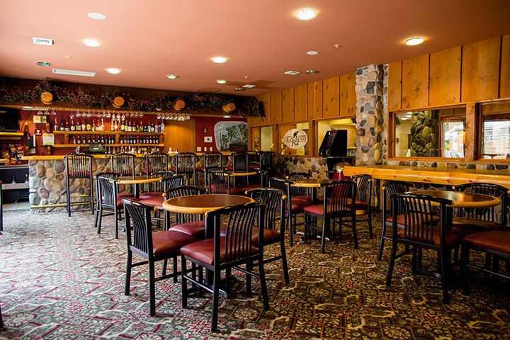 Interior of a restaurant/bar with tables and chairs, a bar with stools, wood paneling, and a patterned carpet.