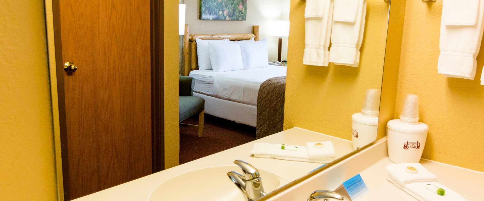 Bathroom interior with a glimpse of a bedroom through an open doorway. The mirror reflects the sink and towels.
