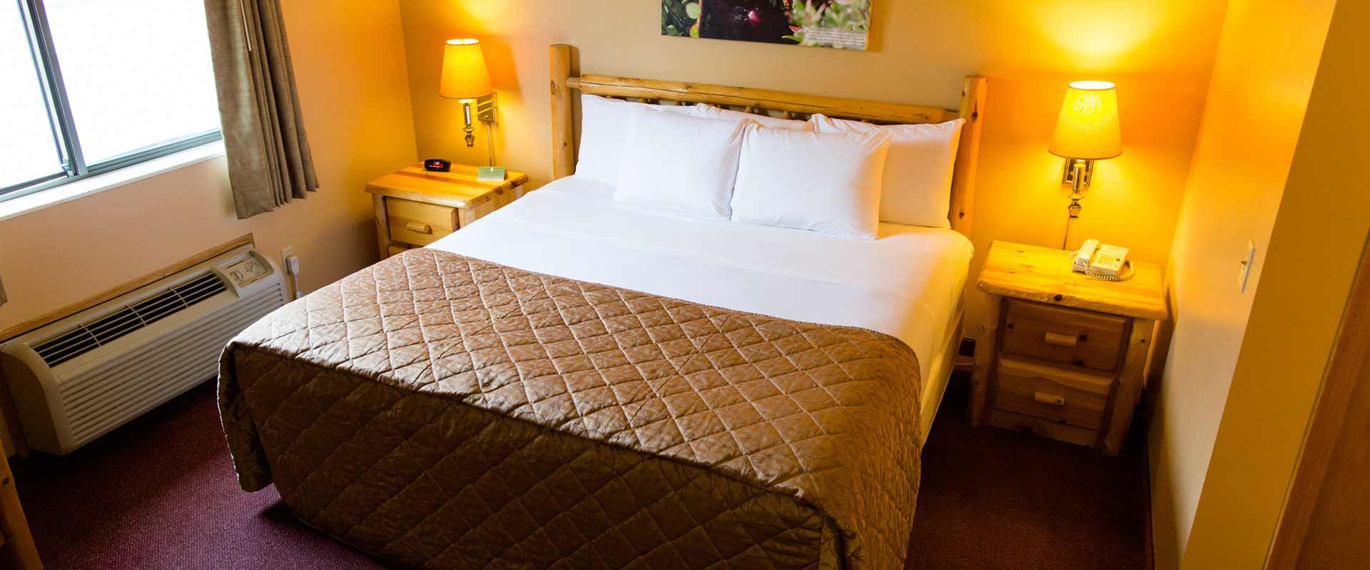 A hotel room with a bed, nightstands, lamps, and a window with an air conditioning unit below.