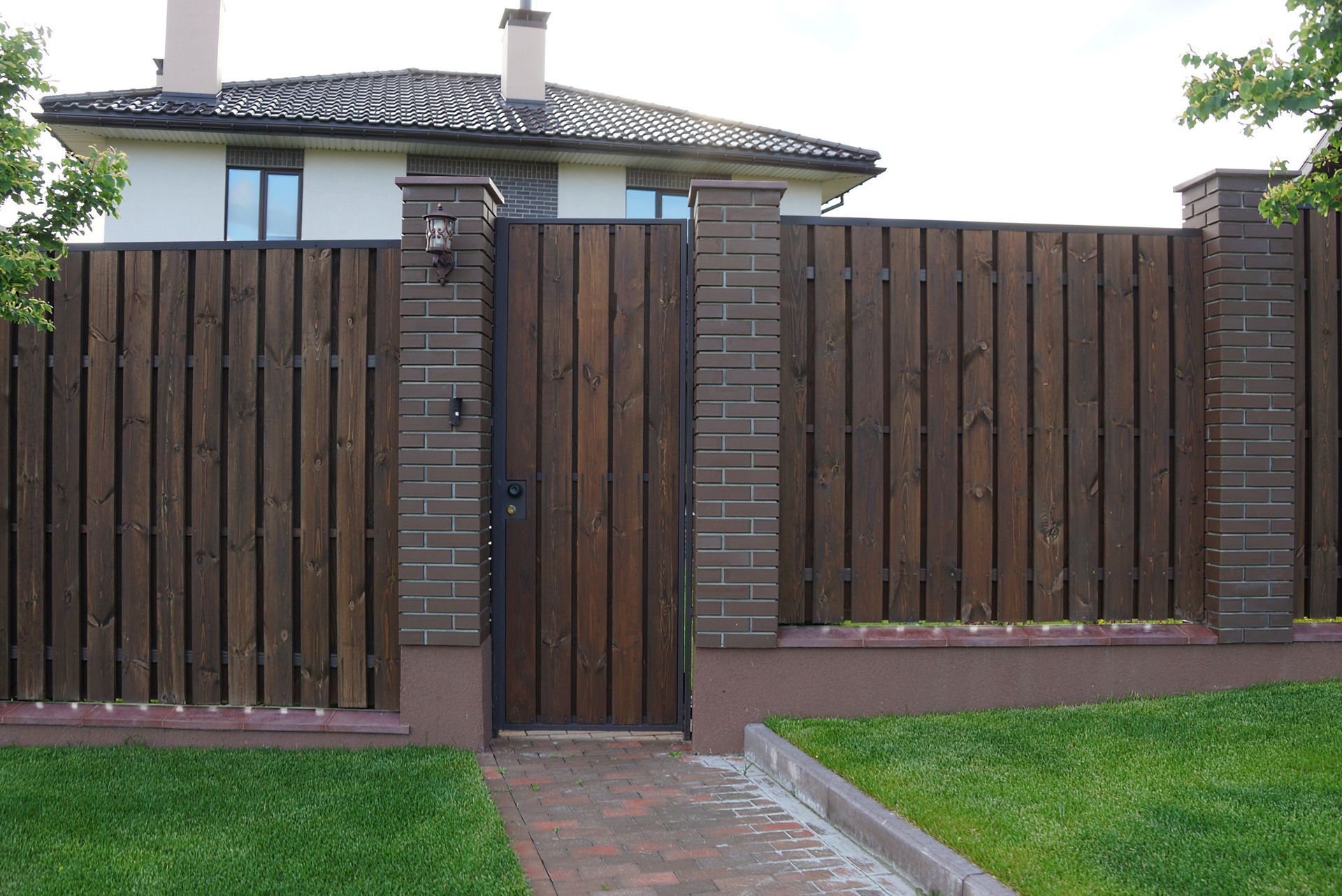 Modern house behind a tall wooden fence with a narrow gate and brick posts, bordered by grass and a paved path