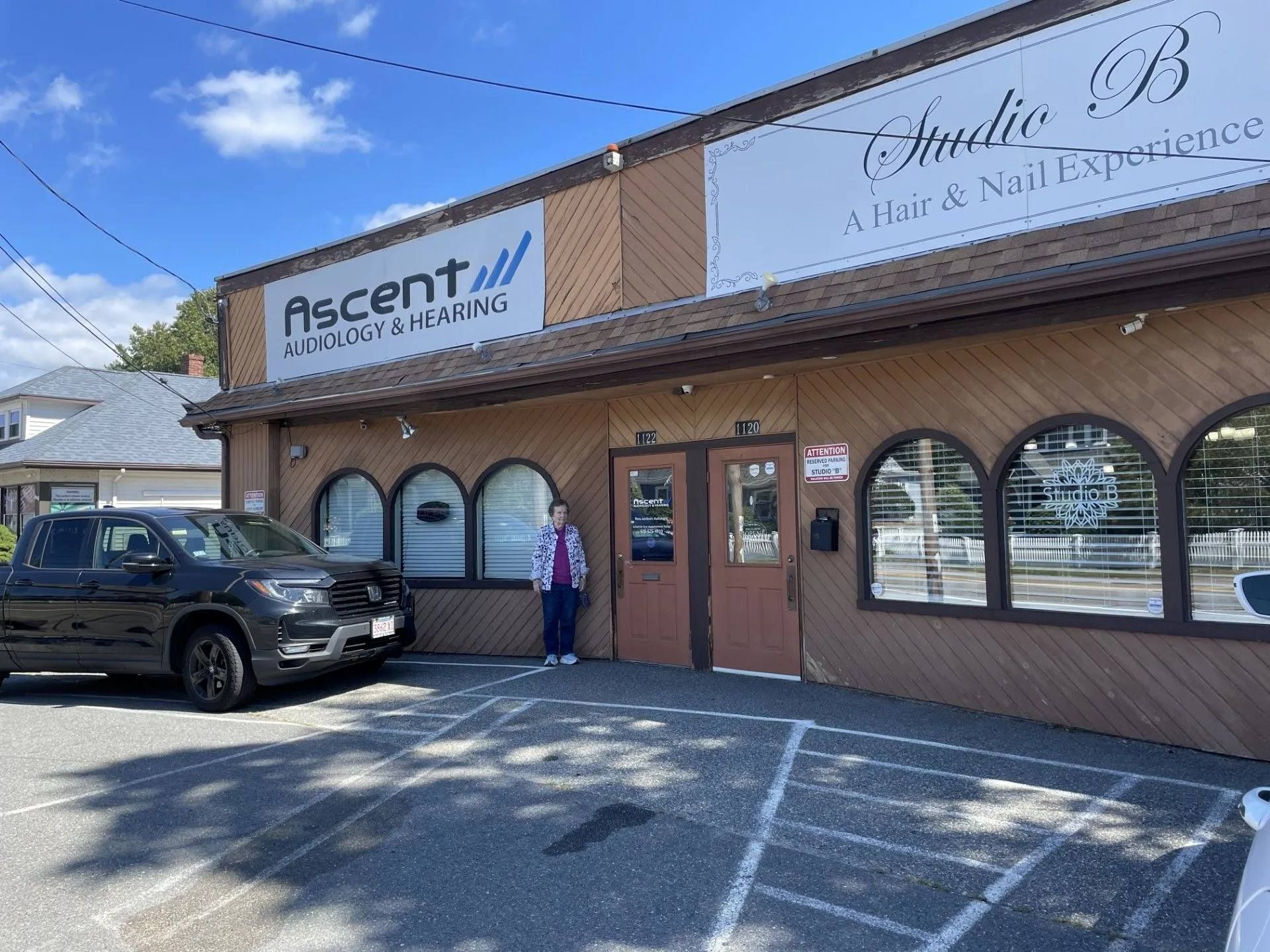 Storefronts in a strip mall with a parked black SUV and one person standing by the entrance