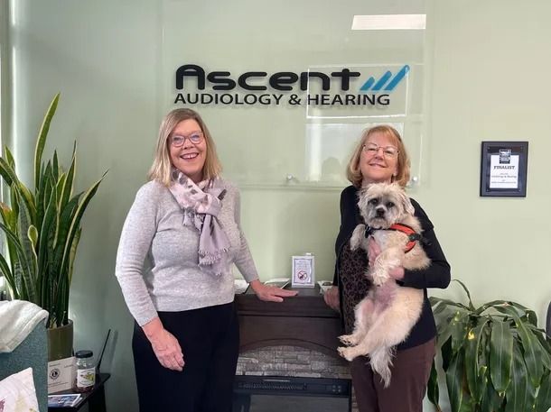 Two women with a dog in front of the Ascent Audiology & Hearing sign