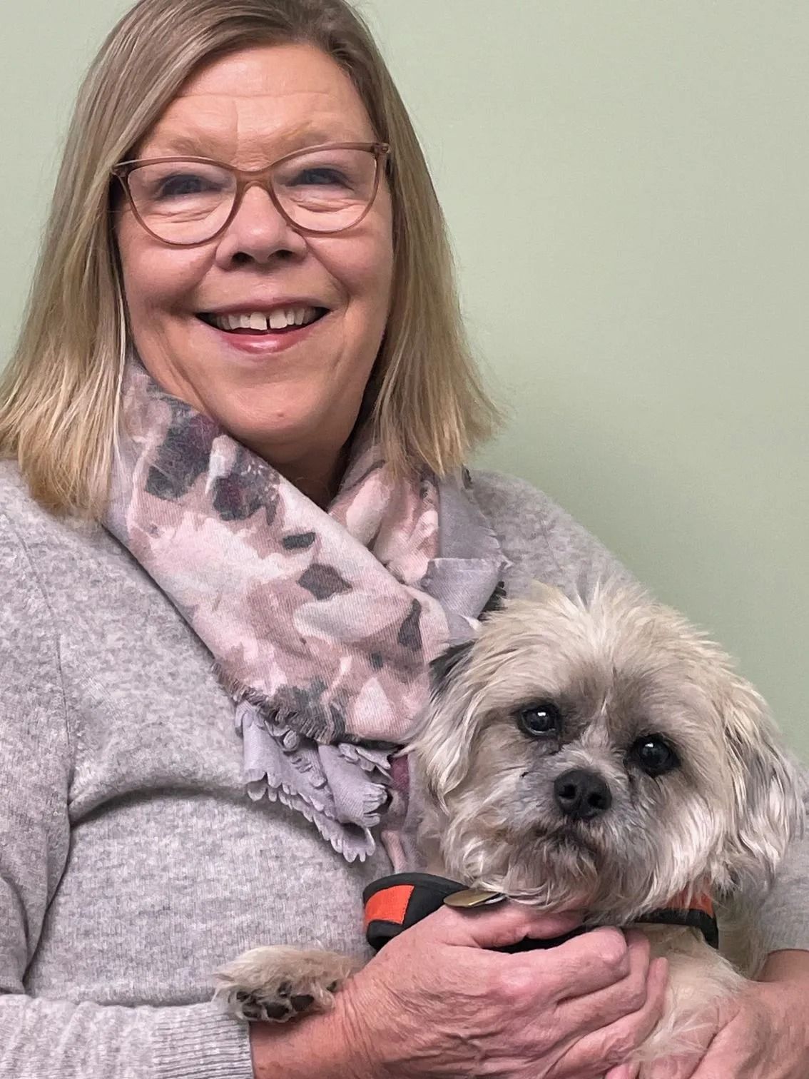 Woman smiling and holding a small shaggy dog, wearing glasses, scarf, and a gray sweater
