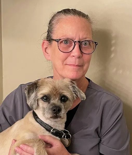 Woman holding a small tan dog with a black collar, both facing the camera against a beige wall.