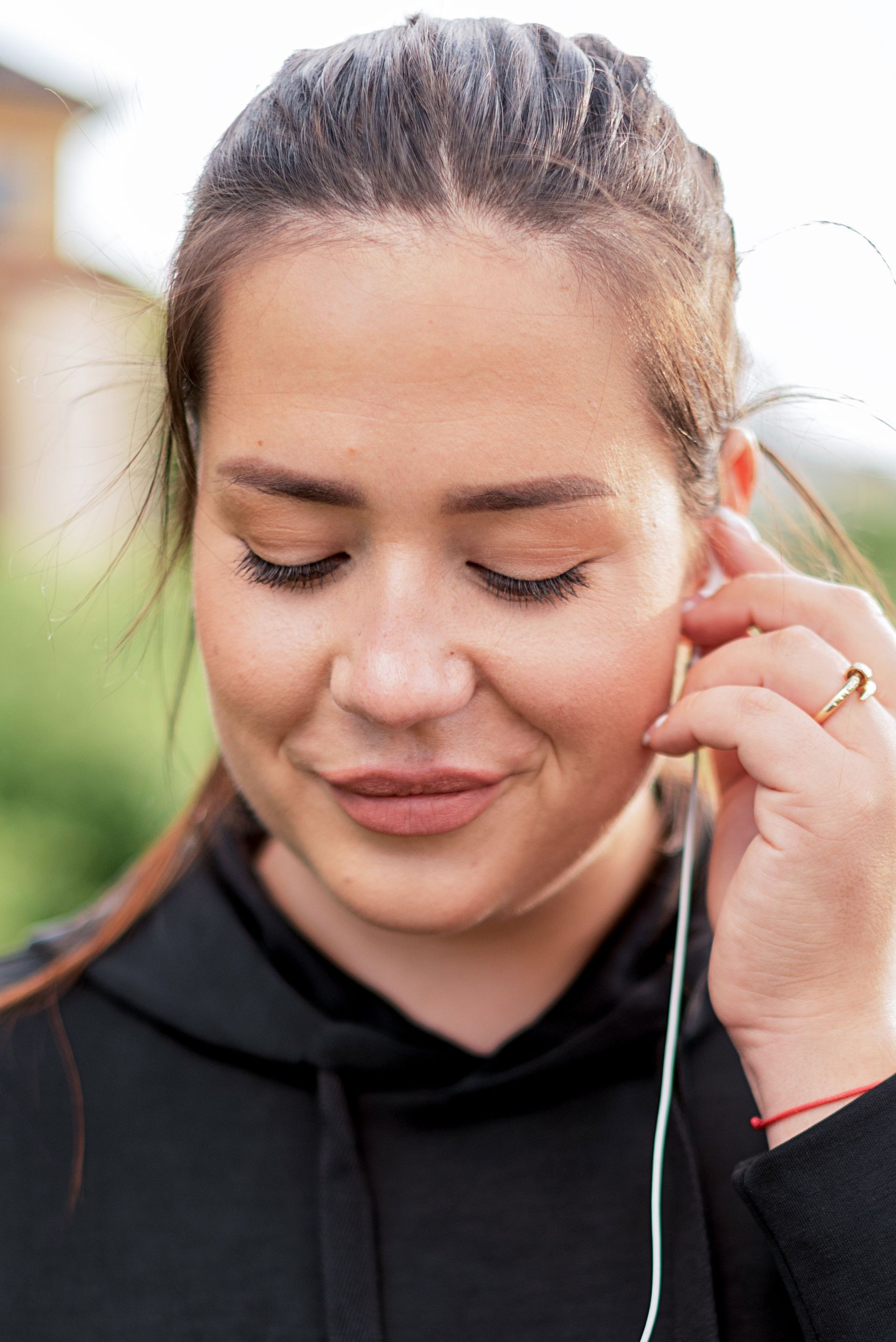 Woman outdoors wearing black hoodie, listening to music with earphones and eyes closed.