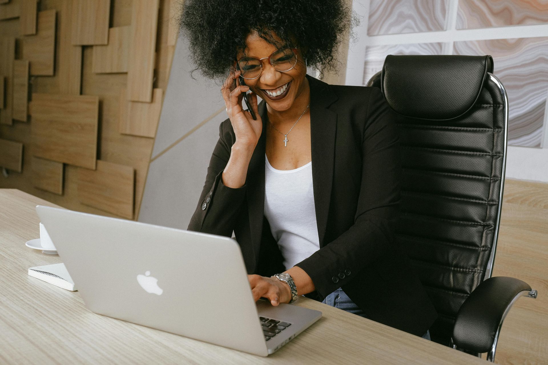 A woman is sitting at a desk using a laptop and talking on a cell phone.