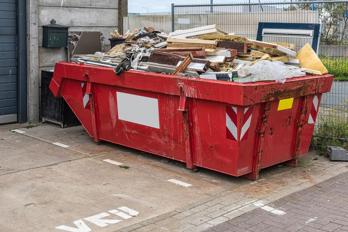A red metal waste container, filled with home renovation debris, stands outside a property.