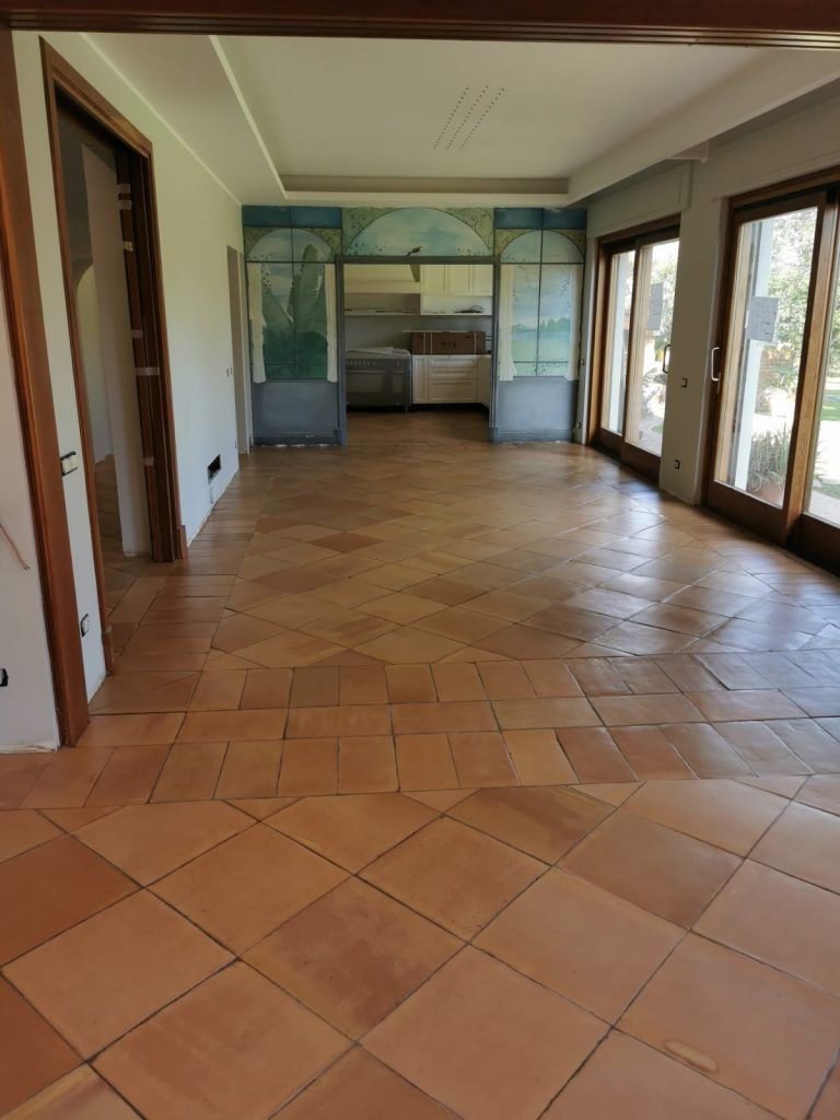 Stanza vuota e luminosa con pavimento in piastrelle di terracotta, finestre con infissi in legno e una nicchia dipinta in fondo.
