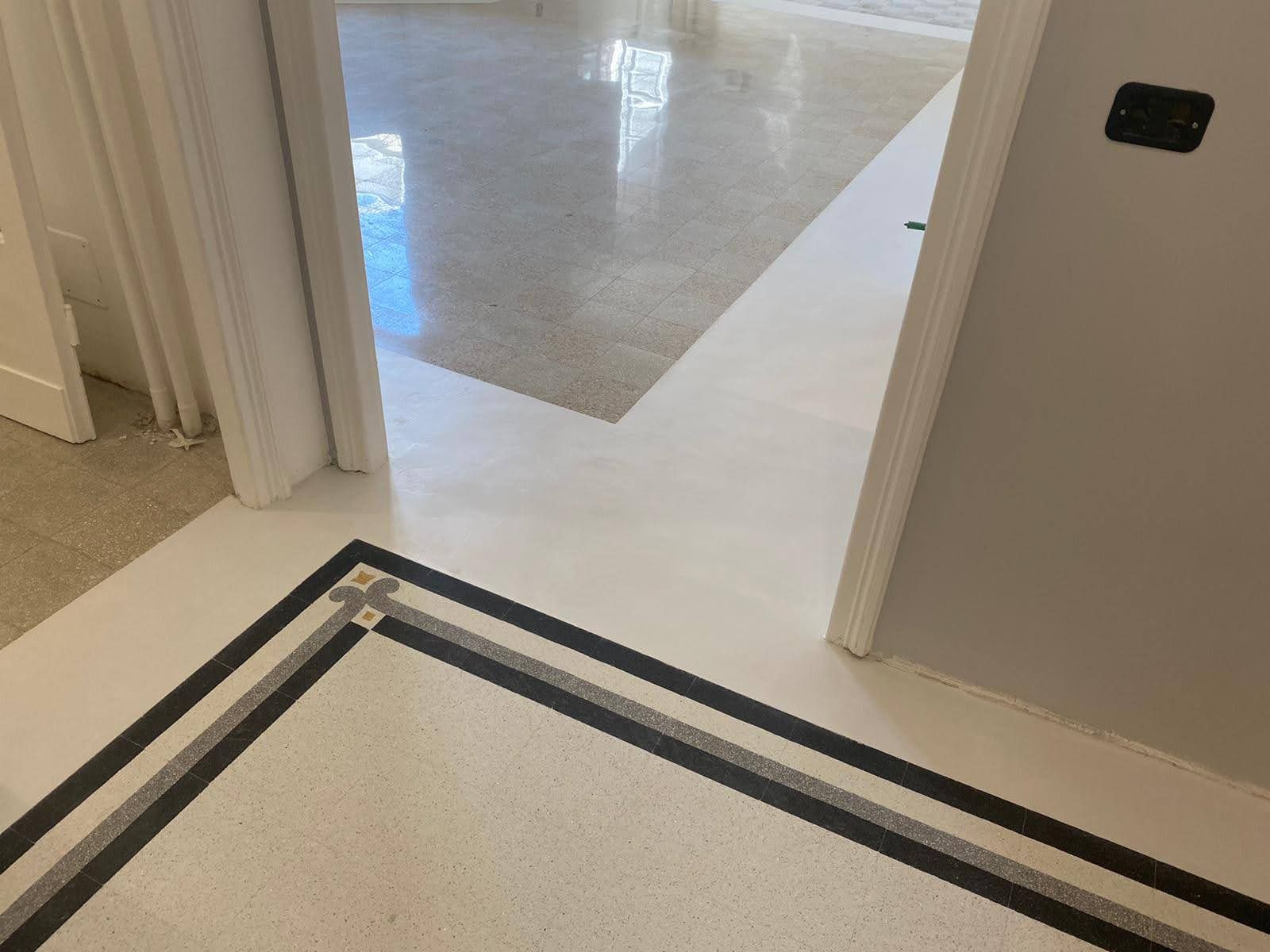Ingresso con pavimento piastrellato bordato di nero che conduce a una stanza luminosa con moquette grigia e pareti bianche.