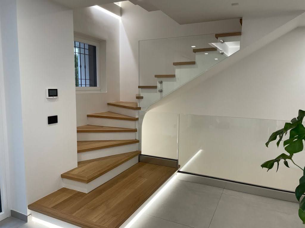 Scala interna moderna con gradini in legno, pareti bianche e ringhiera in vetro in un corridoio luminoso.