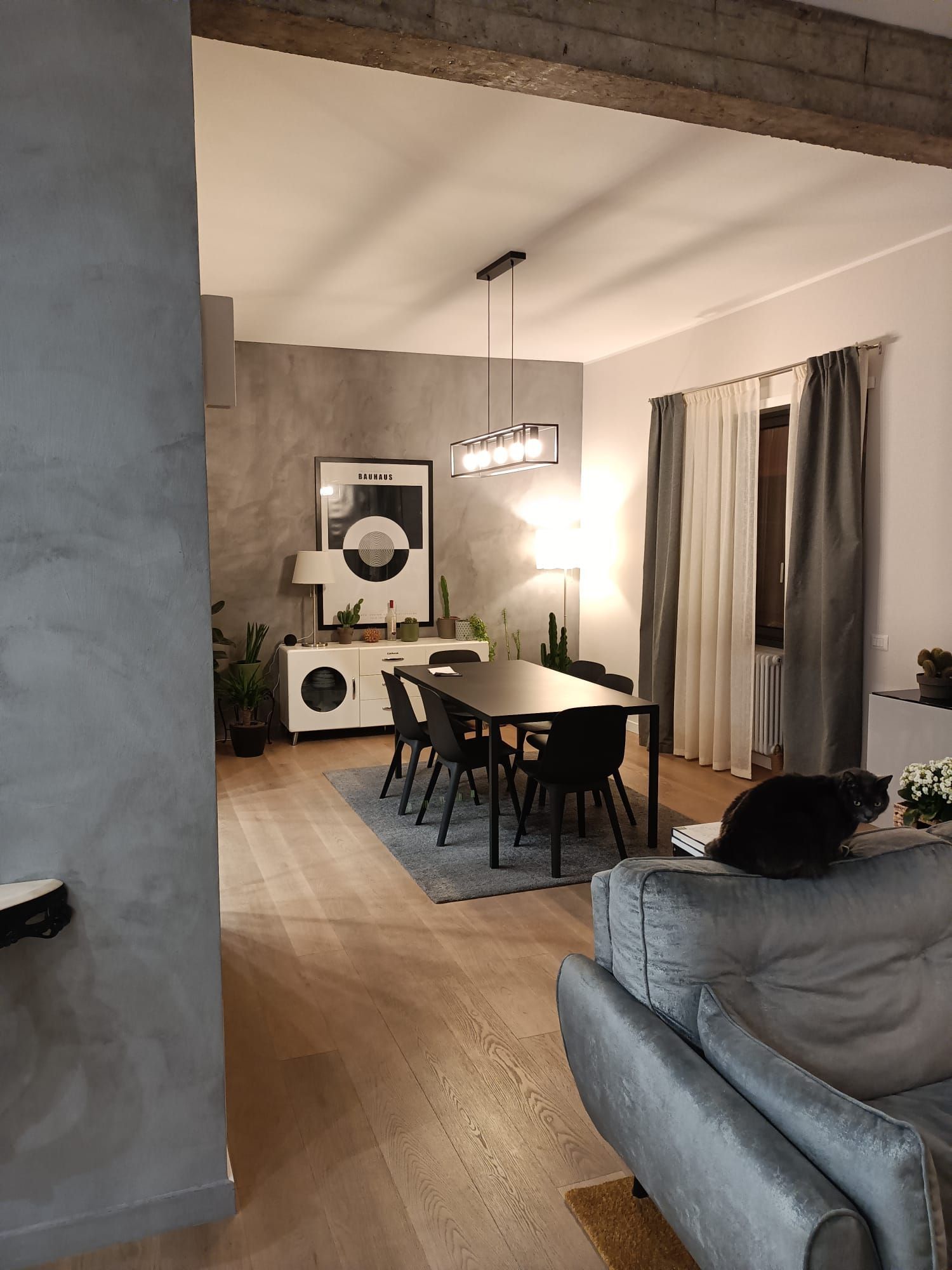 Soggiorno-sala da pranzo moderno con divano grigio, tavolo da pranzo scuro, lampada a sospensione e tende a una finestra.