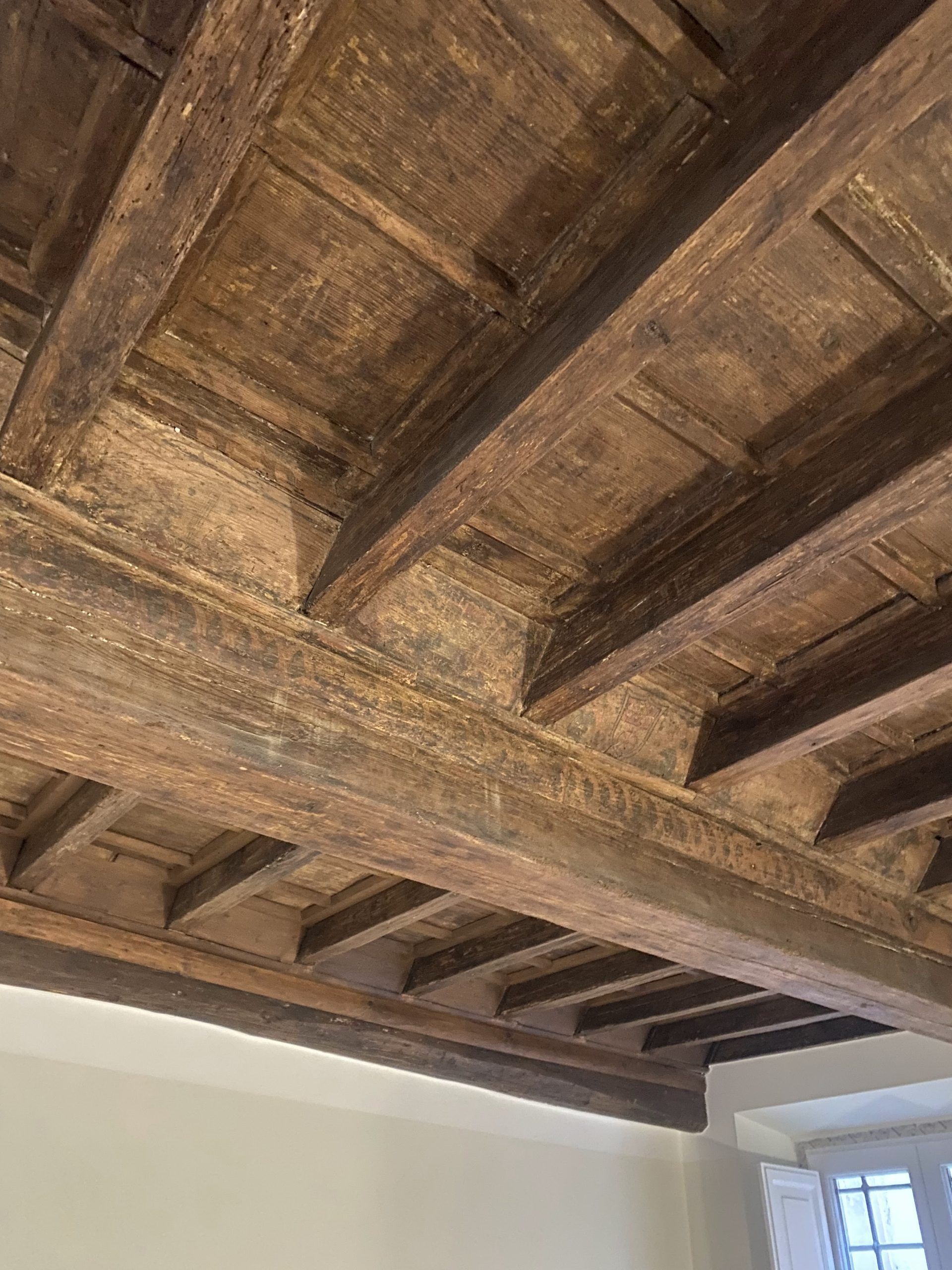 Primo piano di un soffitto rustico con travi in legno, finestra e angolo di parete bianca sottostante.