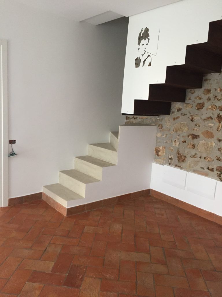 Scala interna con gradini beige, pavimento in piastrelle rosse e una parete bianca accanto a una parete decorativa in pietra.